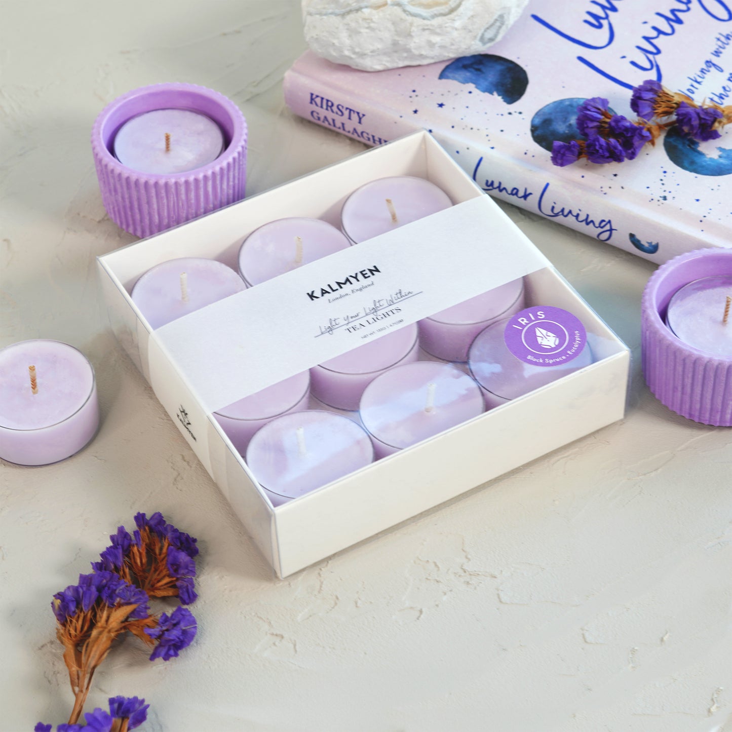 Image of an Iris tea light surrounded by loose tea lights and a purple tealight cup, accompanied by natural flowers and a lilac book in the background.