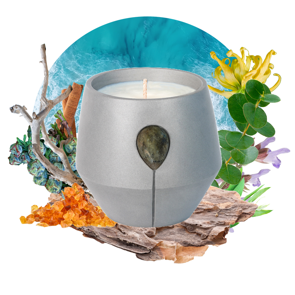 Image of Luna Coast candle with its scent collage at the back - the text reads: Stand at the edge of the shoreline the ocean gently opening your senses. Enriched with calming sweet woods and a resinous base of minty eucalyptus. This scent grounds and balances. Labradorite glistening in the silvery grey vessel.