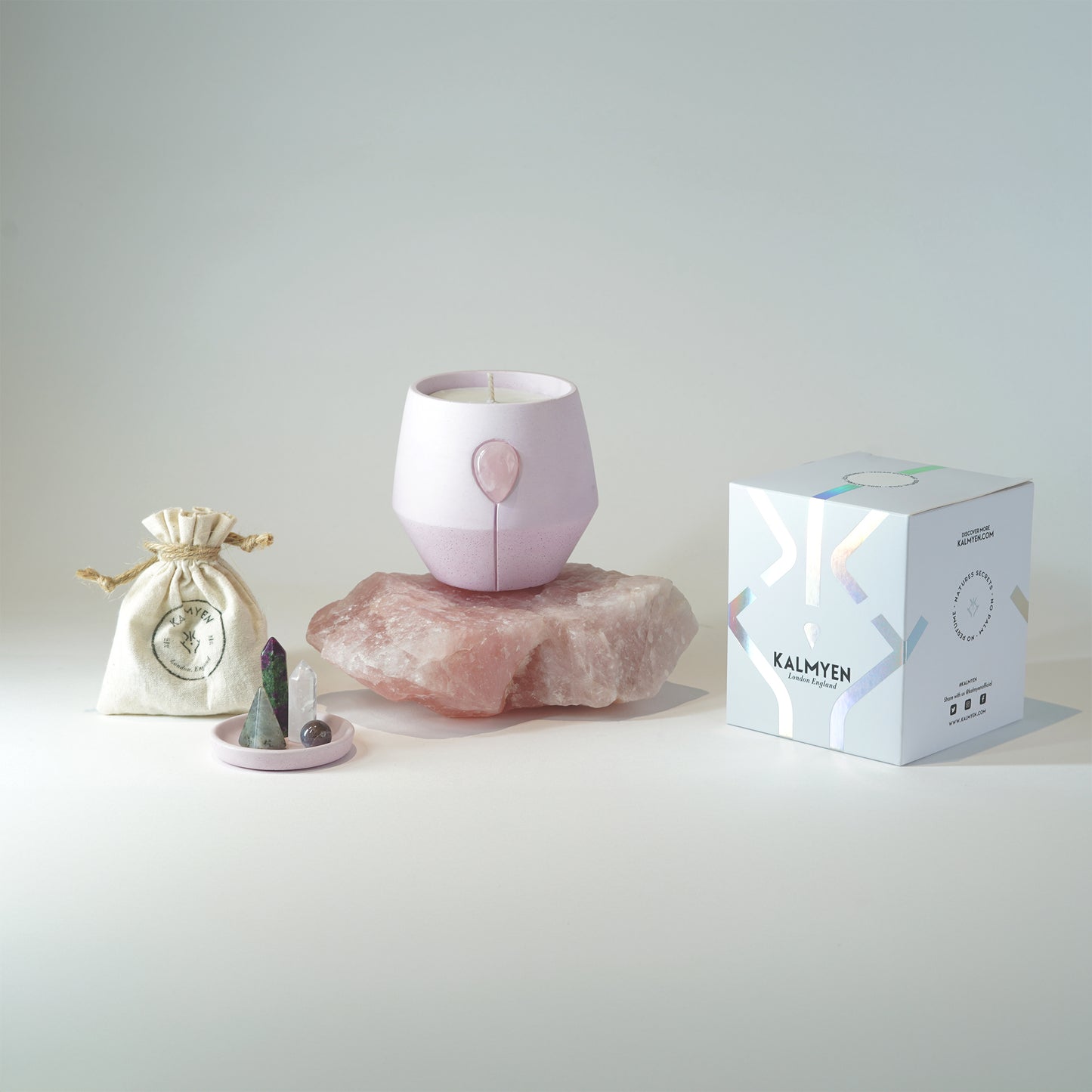 Kalmyen Aurora Luna candle resting on a raw large rose quartz crystal, Luna box on the right side, and a cotton pouch with a mini crystal tray set with four crystals on the left side.