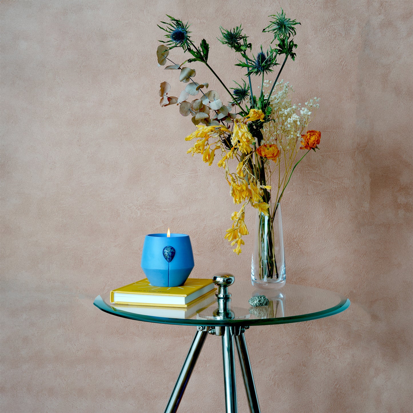 Petrichore Luna candle burning sat ontop of a yellow book on a glass table with a vase of natural dried flowers. 