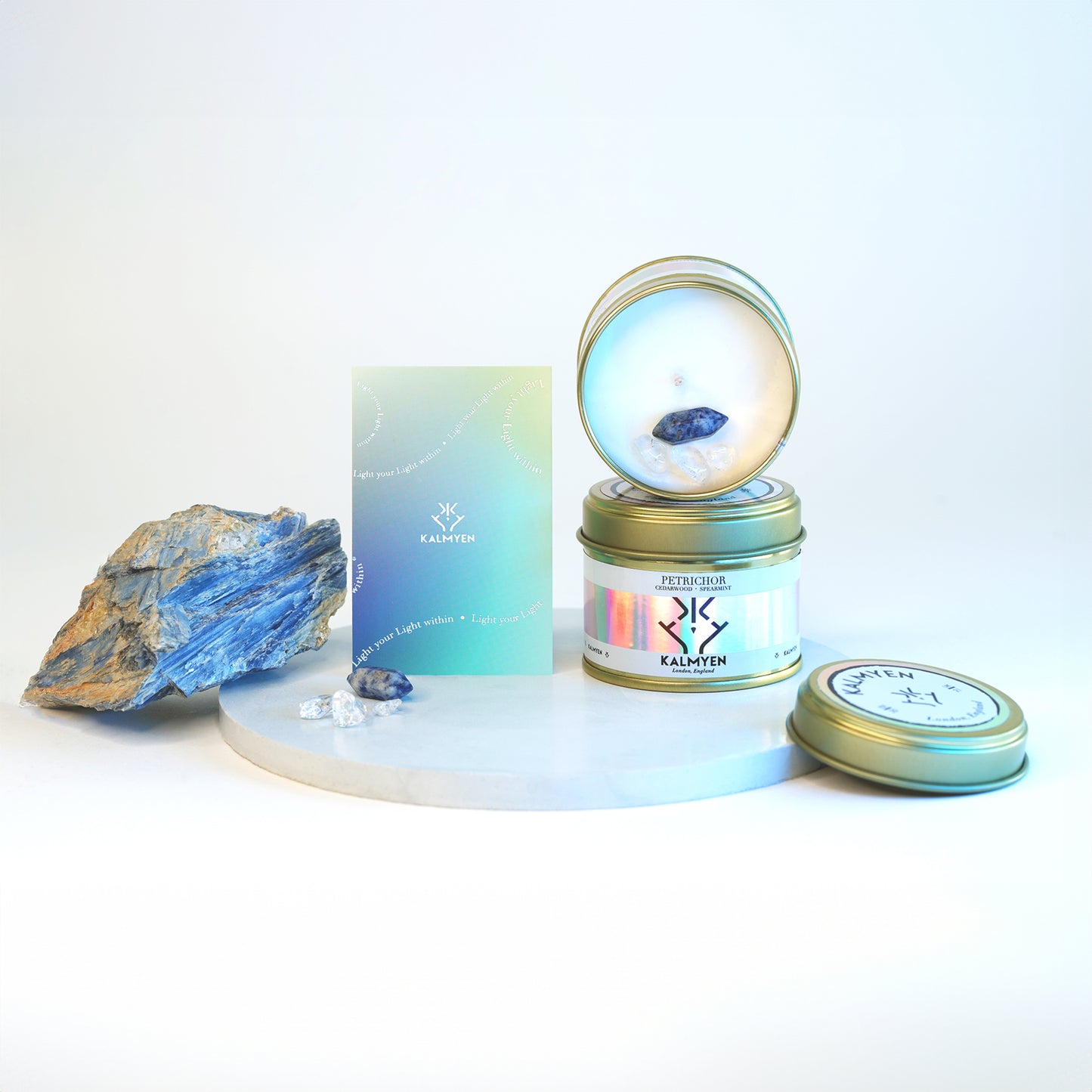 Image showing two Petrichore keepsake tin candles stacked on top of each other the top one is open showing the inside of the keepsake candle embedded with its Blue Spot Jasper mini crystal and clear quatz chips, the candles are diplayed on a blue disc with a raw crystal on the left, a gradient coloured scent card stands next to the raw crystal. 