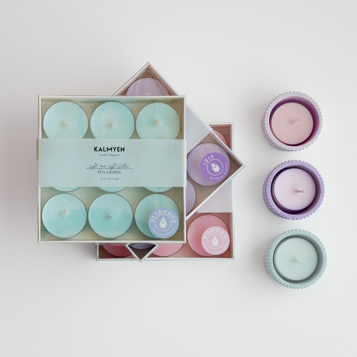 Stacked Kalmyen tealight boxes with a row of three tea light cups on the right side. Minty blue petrichor, lilac iris, and pastel pink aurora tealights and cups are displayed.