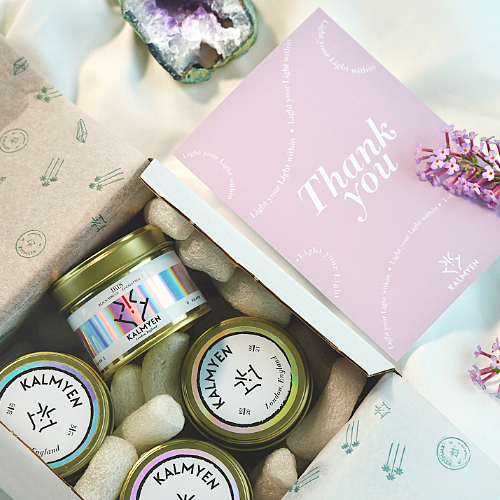 Image showing how keepsake candles are packed with a Kalmyen "thank you" card with a lilac colour and natural lilac flower set to the right side of the image, there are four tins inside the box packed with natural packing peanuts and wrapped in Kalmyen tissue paper. 