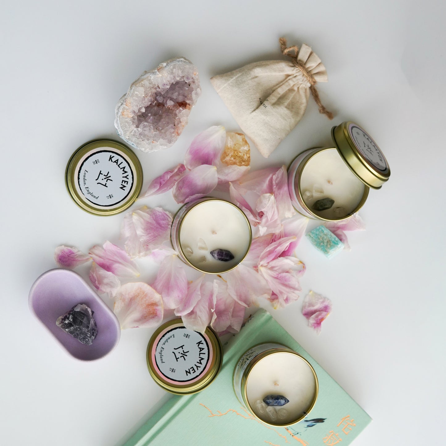 Top-down view of Kalmyen Keepsake tins surrounded by pink peony petals and raw amethyst crystals, with citrine and a cotton pouch. A mini oval tray with fluorite crystal is on the left, and a light green book with a petrichor keepsake tin is at the bottom.