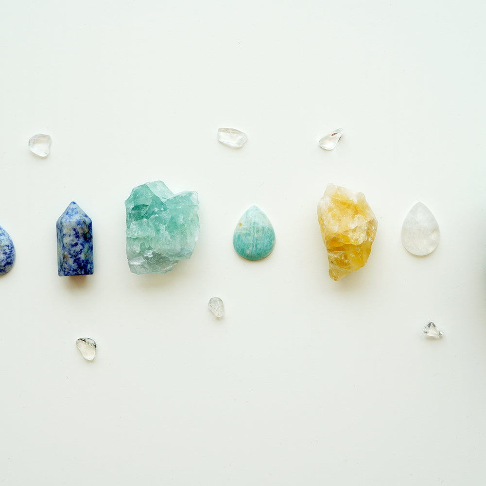 Line of crystals from raw to polished: Blue spot jasper tear drops, mini point, and amazonite nugget; tear drop shaped amazonite crystal; citrine nugget and clear quartz tear drop; celestite rock on the right side. Mini tumbled stones of clear quartz scattered throughout.