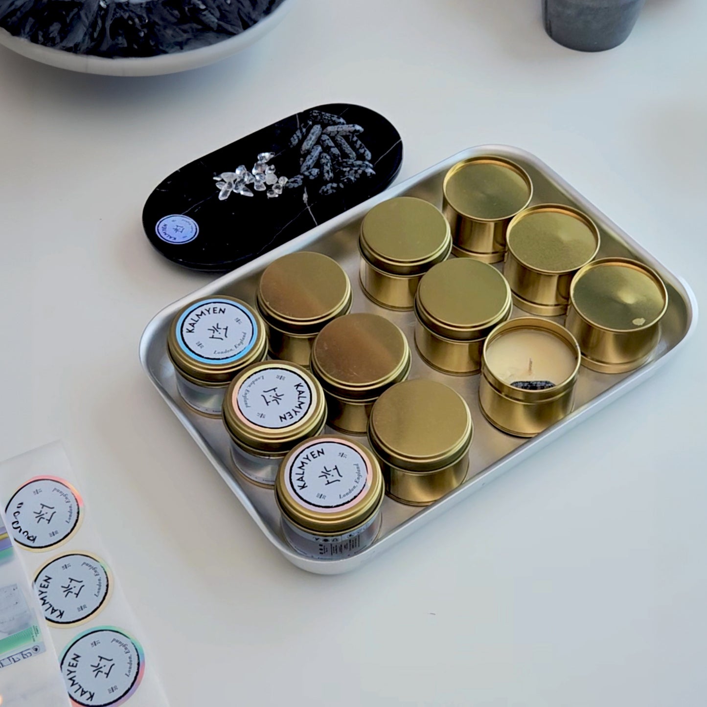 Kalmyen Keepsake tins being embedded with crystals and labeled. A marble tray is positioned behind a tray of tins, with crystal gemstones ready to be added to the wax. The image captures the meticulous process of crafting each candle with care and precision.